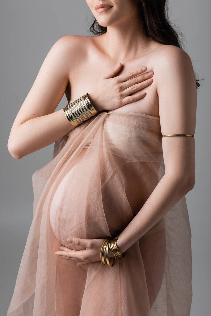 ořezaný pohled na stylovou a elegantní těhotnou ženu pózující ve zlatých doplňcích a průhledné šifonové závěsy izolované na šedém pozadí, koncepce mateřské módy, budoucí matka s břichem - Fotografie, Obrázek