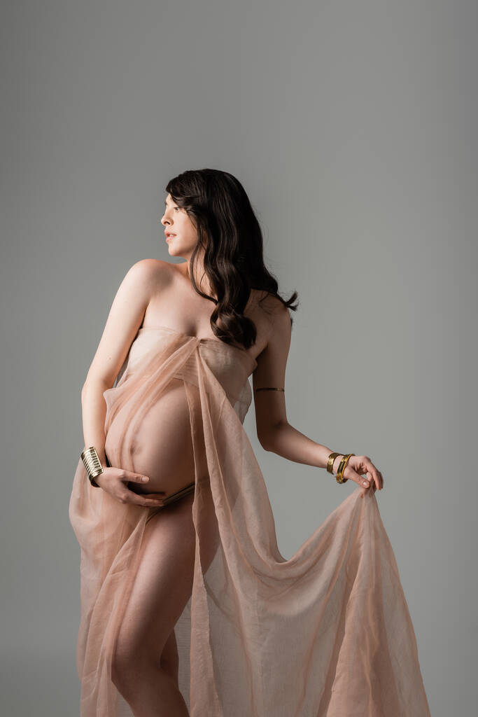 půvabná a těhotná žena s vlnitou brunetkou vlasy pózuje v průhledném šifonu závěsy a dotýká břicho izolované na šedém pozadí, mateřství módní koncept, budoucí matka s břichem  - Fotografie, Obrázek
