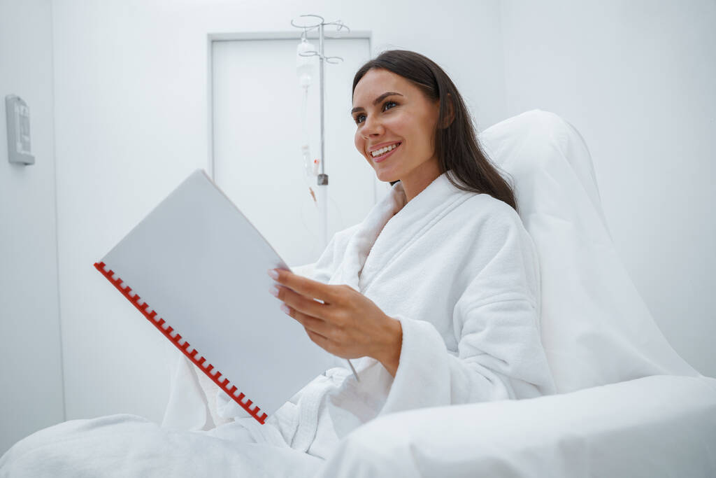 Kobieta w białym szlafroku czyta magazyn podczas siedzenia w luksusowym centrum wellness - Zdjęcie, obraz