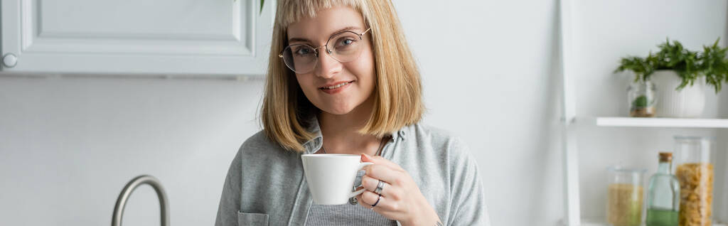 felice giovane donna con i capelli corti e frangia e occhiali in possesso di tazza di caffè del mattino mentre in piedi in abiti casual accanto all'armadio da cucina e pianta in appartamento moderno, banner - Foto, immagini