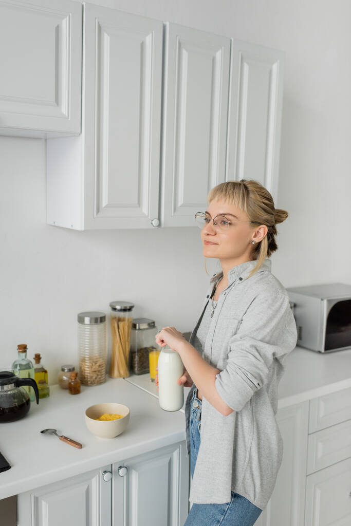 junge Frau mit Brille und kurzen Haaren mit Pony hält Flasche mit Milch in der Nähe Schüssel mit Cornflakes beim Frühstück und steht in lässiger Kleidung neben Küchengeräten zu Hause  - Foto, Bild