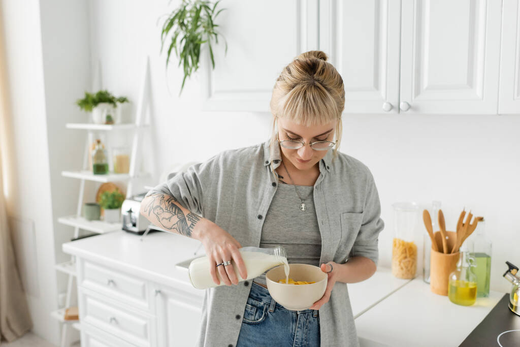 giovane donna tatuata con frangia in occhiali che tiene in bottiglia mentre versa latte fresco in ciotola con cornflakes e prepara la colazione mentre si trova in piedi in abiti casual in cucina moderna  - Foto, immagini