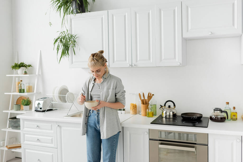 young woman with bangs in eyeglasses holding bowl with cornflakes and spoon while standing in casual grey clothes and denim jeans next to kitchen appliances in blurred white kitchen at home  - Photo, Image