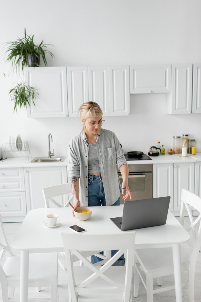 Freelancer tatuado com franja e óculos segurando tigela com flocos de milho enquanto toma café da manhã e olha para laptop perto de smartphone e xícara de café no pires e mesa na cozinha moderna  - Foto, Imagem