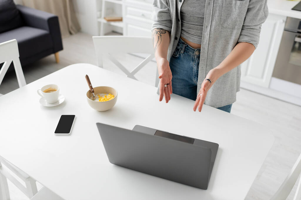 Akıllı telefonun yanında elinde dövmeli genç bir kadın, boş ekranlı bir laptop, mısır gevrekli bir kase, modern mutfağın masasındaki beyaz çay tabağında bir fincan kahve ve bir fincan kahve. - Fotoğraf, Görsel