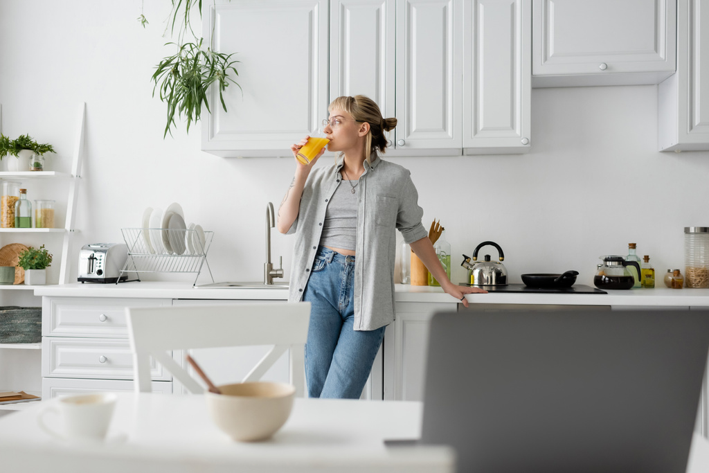 tattooed woman in eyeglasses drinking orange juice and standing near  kitchen worktop next to desk with devices, bowl with cornflakes and cup of coffee with saucer at home, freelance lifestyle  - Photo, Image