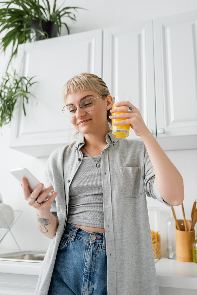 potetovaná a šťastná žena s ofinou a brýlemi držící sklenici pomerančové šťávy a používající smartphone, zatímco stojí v blízkosti čistých jídel a rozmazaných zelených rostlin v moderním bytě  - Fotografie, Obrázek