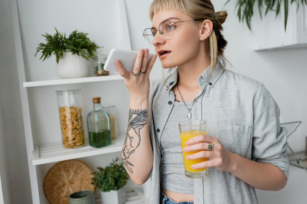 tatuoitu nuori nainen otsatukka ja silmälasit tilalla lasi appelsiinimehua ja tallennus ääniviestin älypuhelimeen, seisoo lähellä hämärtynyt vihreitä kasveja ja teline moderni valkoinen keittiö   - Valokuva, kuva