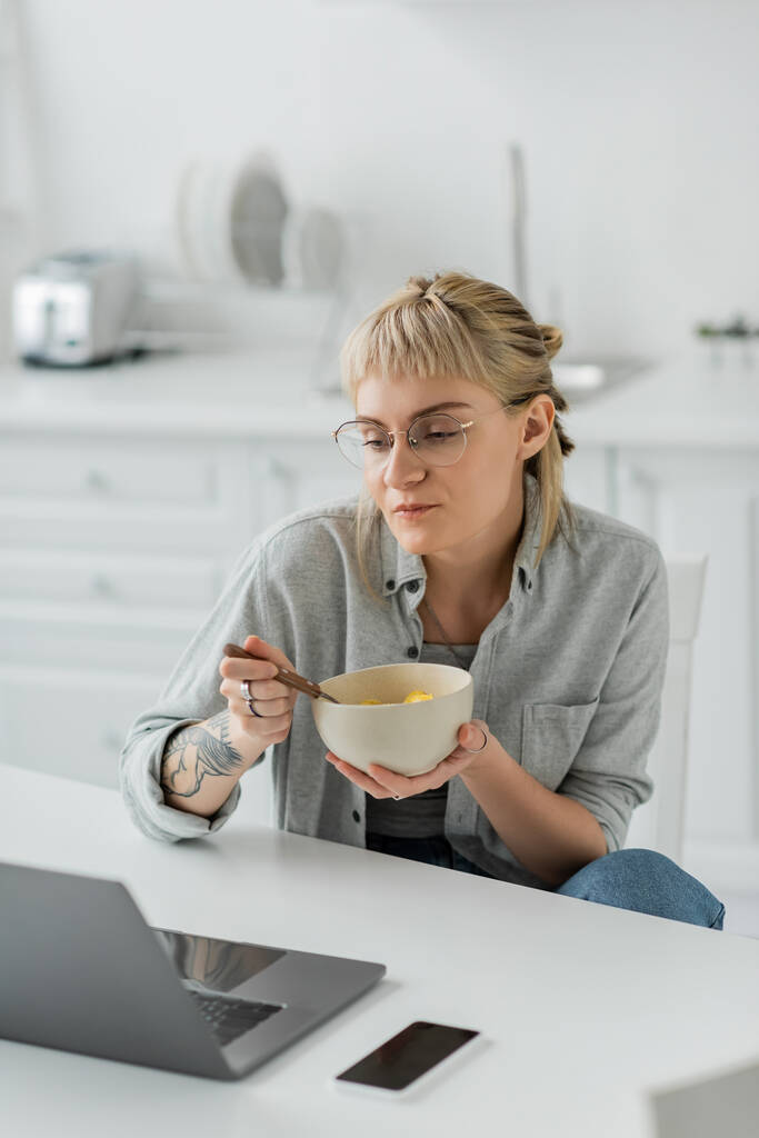 молодая женщина с челкой и татуировкой на руке едят кукурузные хлопья на завтрак, глядя на ноутбук рядом со смартфоном с чистым экраном на столе в современной кухне, фрилансер, работа из дома  - Фото, изображение