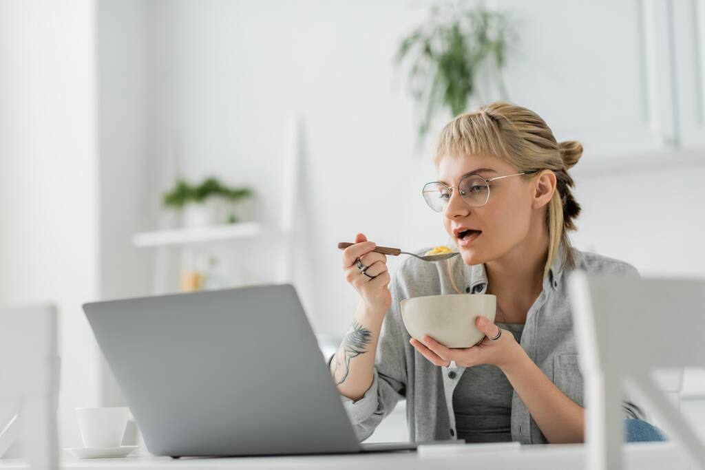 νεαρή γυναίκα με αφέλειες και τατουάζ στο χέρι τρώει κορν φλέικς για πρωινό, ενώ χρησιμοποιώντας το φορητό υπολογιστή κοντά στο smartphone και το φλιτζάνι του καφέ στο τραπέζι στη σύγχρονη κουζίνα, ελεύθερος επαγγελματίας  - Φωτογραφία, εικόνα