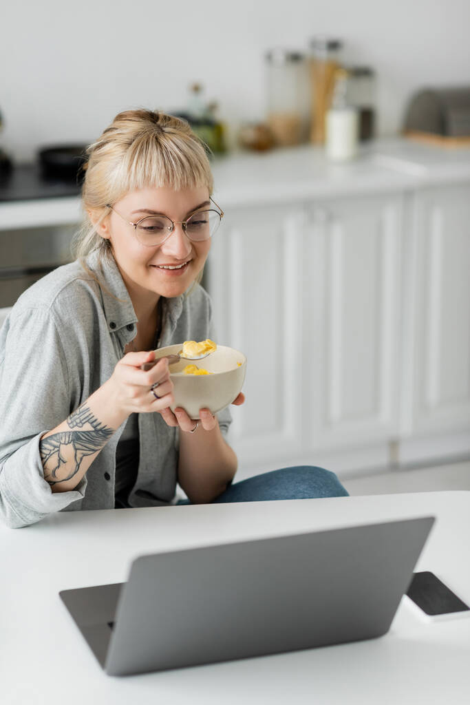 χαρούμενη νεαρή γυναίκα με κοντά μαλλιά, αφέλειες και τατουάζ στο χέρι τρώει κορν φλέικς για πρωινό, ενώ χρησιμοποιώντας το φορητό υπολογιστή κοντά στο smartphone με λευκή οθόνη στο τραπέζι και χαμογελώντας στη σύγχρονη κουζίνα, ελεύθερος επαγγελματίας  - Φωτογραφία, εικόνα