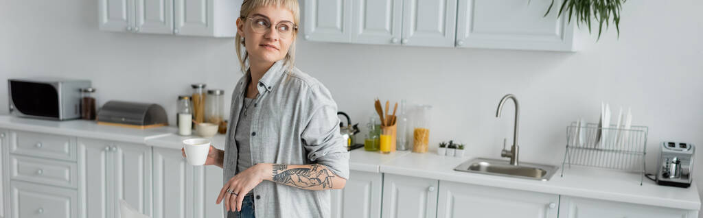 νεαρή γυναίκα με γυαλιά ηλίου με τατουάζ στο χέρι και αφέλειες κρατώντας φλιτζάνι καφέ και κοιτάζοντας μακριά, ενώ στέκεται στη σύγχρονη κουζίνα με διαφορετικές συσκευές και λευκά ντουλάπια, πανό, την πρωινή ώρα  - Φωτογραφία, εικόνα