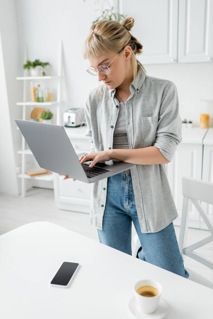 young woman in eyeglasses with short hair and bangs holding laptop near cup of coffee and smartphone with blank screen on white table in white and modern kitchen, remote lifestyle, freelancer - Photo, Image