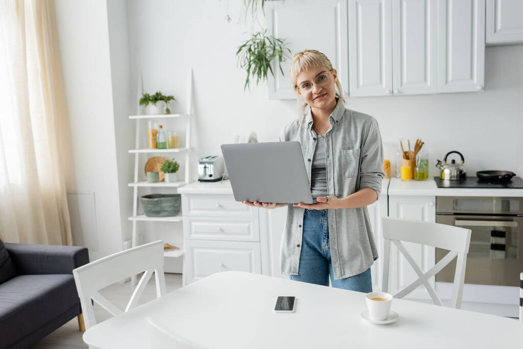 onnellinen nuori nainen silmälasit lyhyet hiukset ja otsatukka tilalla kannettava tietokone ja katsot kameraa lähellä kuppi kahvia ja älypuhelin tyhjä näyttö valkoisella pöydällä valkoinen ja moderni keittiö, freelance - Valokuva, kuva