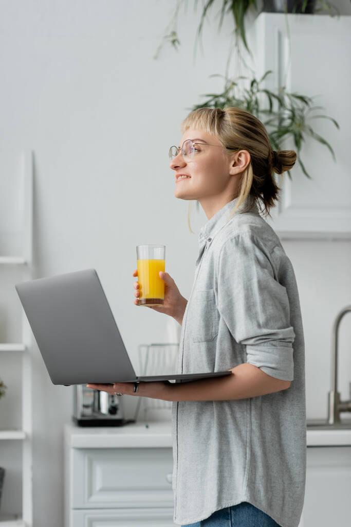 happy young woman in eyeglasses, short hair and bangs holding glass of fresh orange juice and laptop while working from home in white and modern kitchen, blurred background, freelancer - Photo, Image