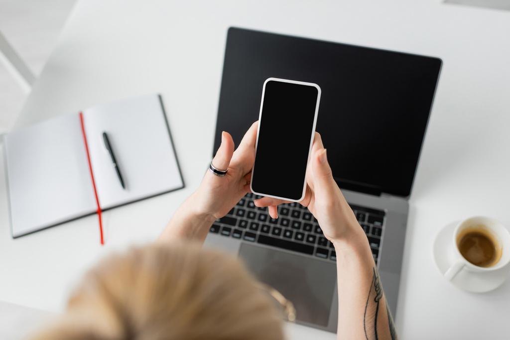 widok z góry wytatuowany kobieta gospodarstwa smartfon z pustym ekranem w pobliżu laptopa, notebook z piórem i filiżanka kawy z talerzykiem na białym stole podczas pracy w domu, freelancer, nowoczesne miejsce pracy  - Zdjęcie, obraz