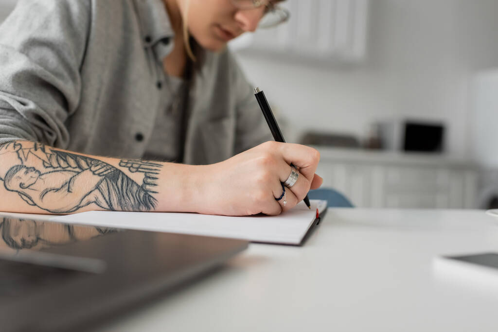 vista recortada de jovem com tatuagem na mão escrevendo em caderno, tomando notas, tendo inspiração enquanto segurando caneta perto de laptop na mesa branca, foreground borrado, trabalho em casa  - Foto, Imagem
