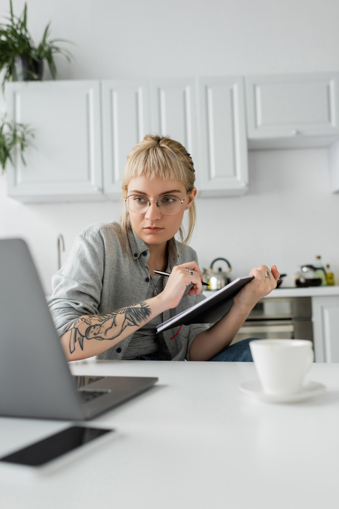молодая женщина с татуировкой на руке и челкой, держащая ноутбук, делая заметки возле смартфона и ноутбука на белом столе, размытый передний план, работа из дома  - Фото, изображение