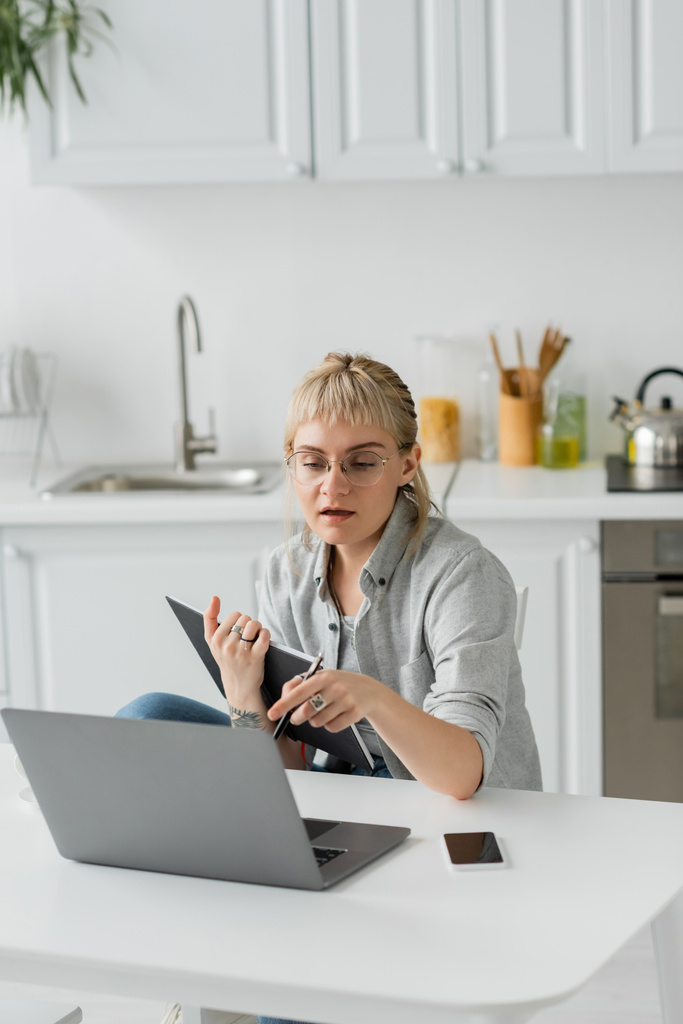 young woman with tattoo on hand and bangs holding notebook, near smartphone with blank screen and looking at laptop on white table, blurred foreground, work from home  - Photo, Image