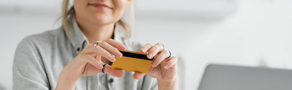cropped view of happy young woman smiling and holding credit card, sitting near blurred laptop in modern apartment, workspace, digital lifestyle,online transactions, technology, banner  - Photo, Image