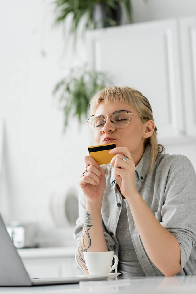 молодая женщина в очках с татуировкой на руке, целующая кредитную карту, сидящая рядом с ноутбуком и чашкой кофе на белом столе, размытый фон, работа из дома, онлайн транзакции, технологии  - Фото, изображение