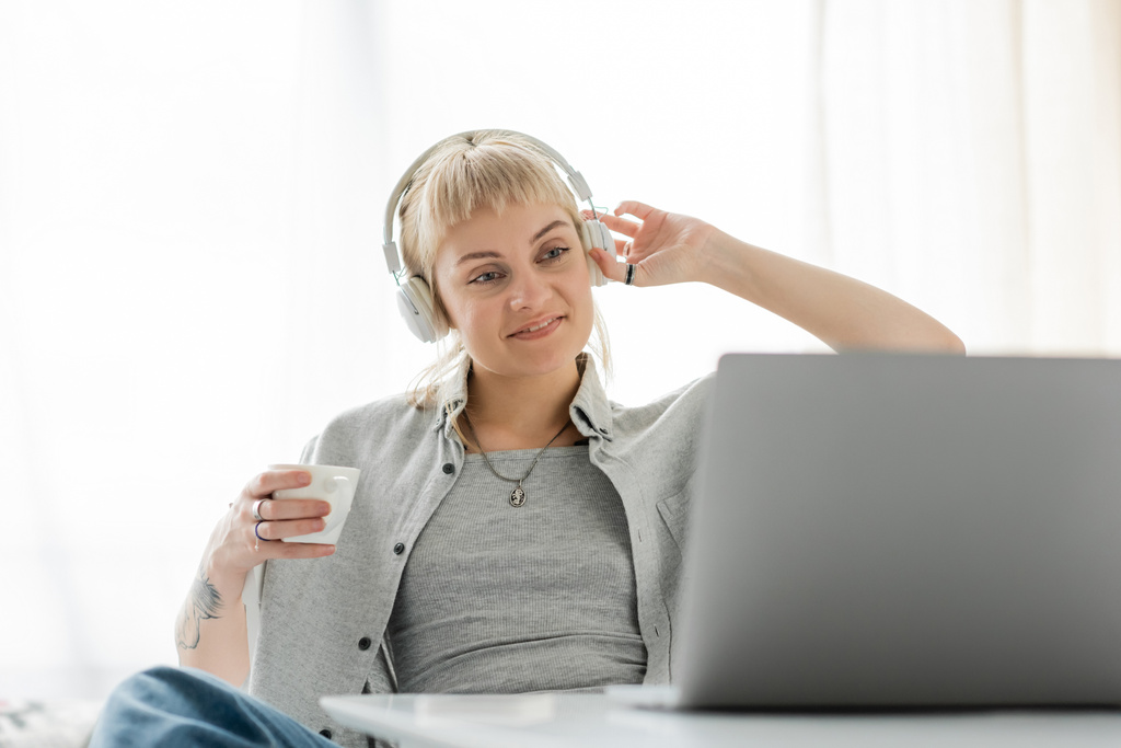glimlachende jonge vrouw met pony en tatoeage op de hand zitten in draadloze koptelefoon en het houden van kopje koffie terwijl het kijken naar laptop, op wazig tafel, freelance, werk van thuis uit - Foto, afbeelding