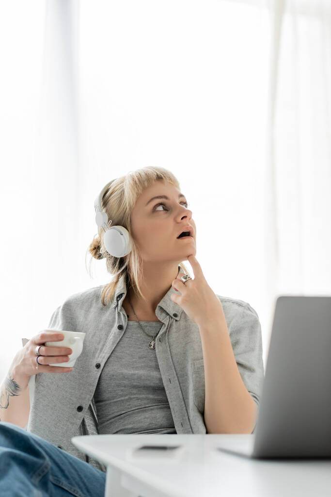σκεπτική νεαρή γυναίκα με ξανθά μαλλιά, αφέλειες και τατουάζ στο χέρι κάθεται σε ασύρματα ακουστικά και κρατώντας φλιτζάνι καφέ κοντά στο laptop και θολή smartphone στο τραπέζι. Ελεύθερος επαγγελματίας. εργασία από το σπίτι - Φωτογραφία, εικόνα