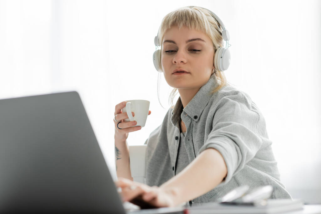 mladý freelancer s blond vlasy, ofina a tetování na ruce sedí v bezdrátových sluchátkách a drží šálek kávy u notebooku a rozmazané notebooky a brýle na stole, práce z domova  - Fotografie, Obrázek