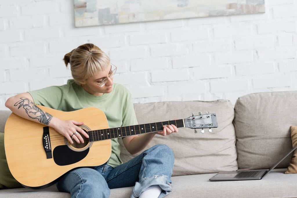 jeune femme dans des lunettes avec frange et tatouage tenant guitare acoustique et apprendre à jouer tout en regardant tutoriel vidéo sur ordinateur portable et assis sur un canapé confortable dans le salon moderne à la maison - Photo, image