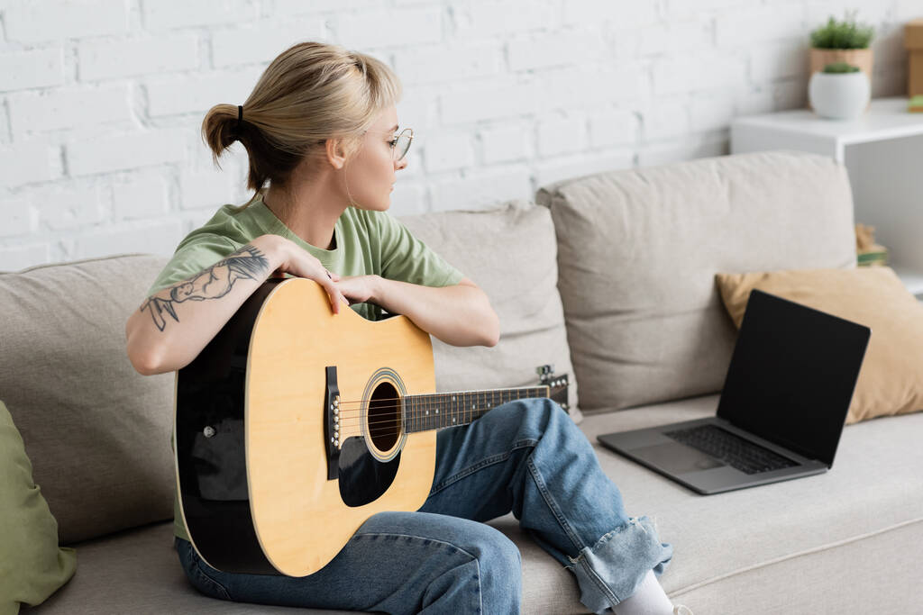 молодая женщина в очках с челкой и татуировкой, держащая акустическую гитару и учится играть, глядя видео-учебник на ноутбуке с чистым экраном и сидя на удобном диване в гостиной - Фото, изображение