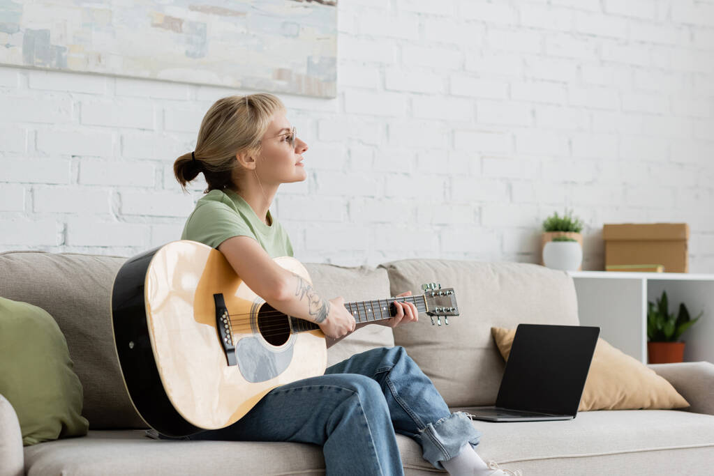 jeune femme dans des lunettes avec frange et tatouage jouer de la guitare acoustique près d'un ordinateur portable avec écran blanc et assis sur un canapé confortable dans le salon, apprendre la musique, le développement des compétences  - Photo, image