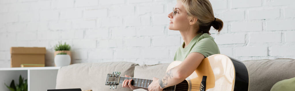 giovane donna in occhiali con frangia e tatuaggio che suona la chitarra acustica e seduta su un comodo divano in soggiorno moderno, apprendimento della musica, sviluppo delle abilità, appassionato di musica, banner  - Foto, immagini