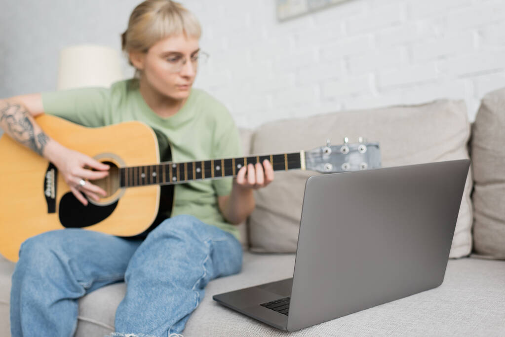 mujer joven borrosa en gafas con flequillo y tatuaje tocando la guitarra acústica y mirando video tutorial en el ordenador portátil mientras está sentado en un cómodo sofá en la sala de estar moderna, aprendizaje virtual  - Foto, imagen