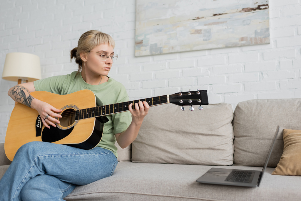 giovane donna in occhiali con frangia e tatuaggio che suona la chitarra acustica mentre cerca video tutorial sul computer portatile e seduta su un comodo divano nel moderno soggiorno di casa, sviluppo delle abilità  - Foto, immagini