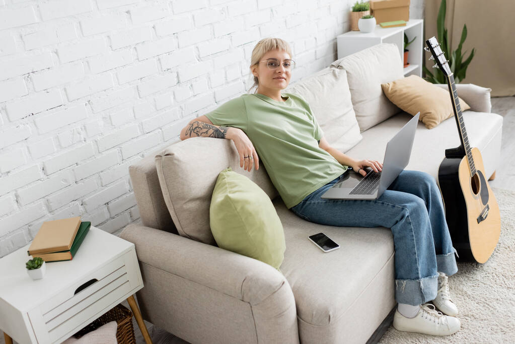 onnellinen tatuoitu nainen, jolla on vaaleat ja lyhyet hiukset, otsatukka ja silmälasit kannettavan tietokoneen avulla istuen mukavalla sohvalla älypuhelimen vieressä ja katsoen kameraa lähellä kitaraa modernissa olohuoneessa  - Valokuva, kuva