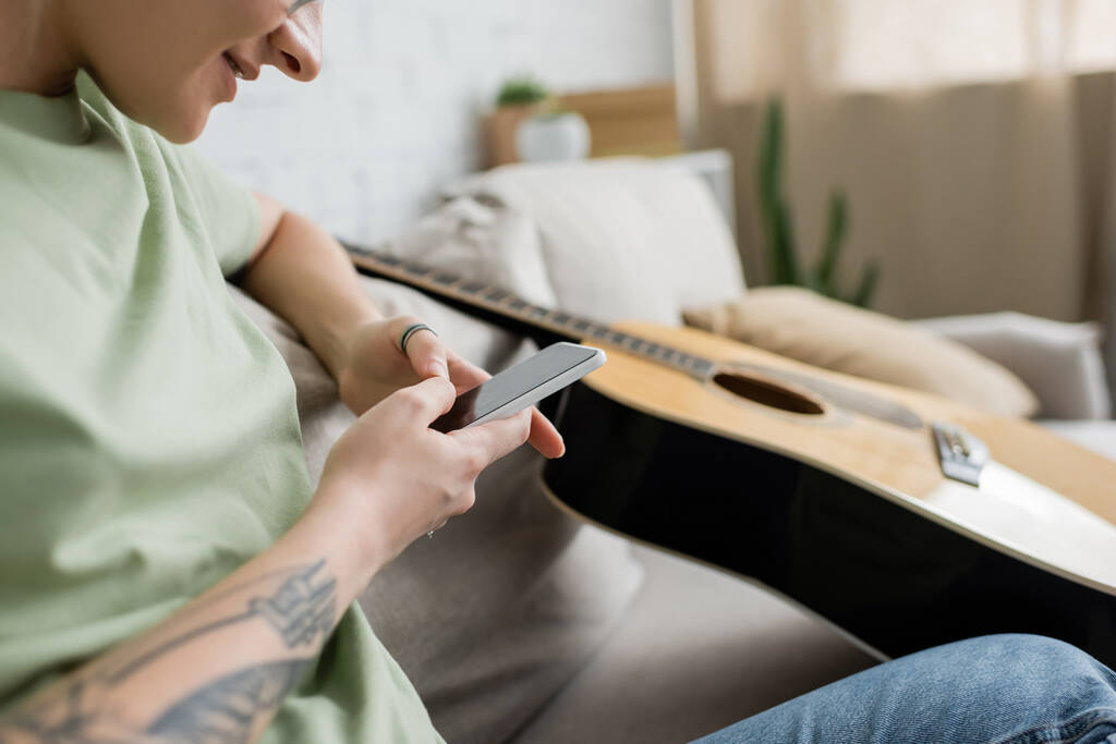 abgeschnittene Ansicht einer glücklichen jungen Frau mit Tätowierung auf der Hand mittels Smartphone, während sie auf einer bequemen Couch neben der Gitarre im modernen Wohnzimmer sitzt, verschwommene Aufnahme  - Foto, Bild