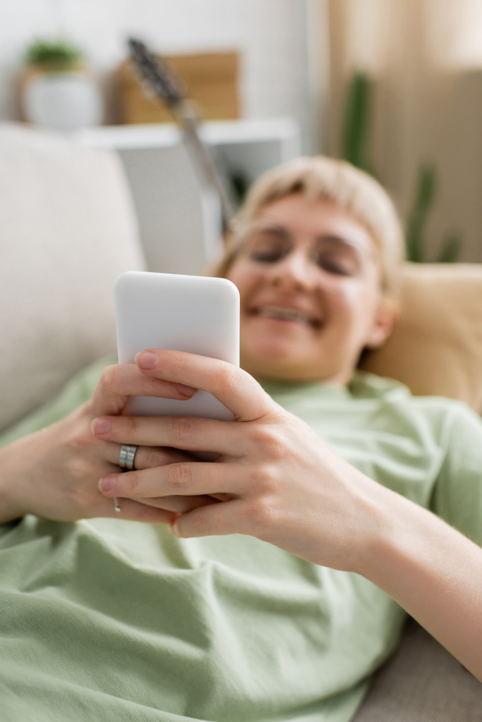 glückliche junge Frau mit Tätowierung, blonden und kurzen Haaren, Pony und Brille mit Smartphone, während sie auf einer bequemen Couch im modernen Wohnzimmer ruht, verschwommener Hintergrund - Foto, Bild