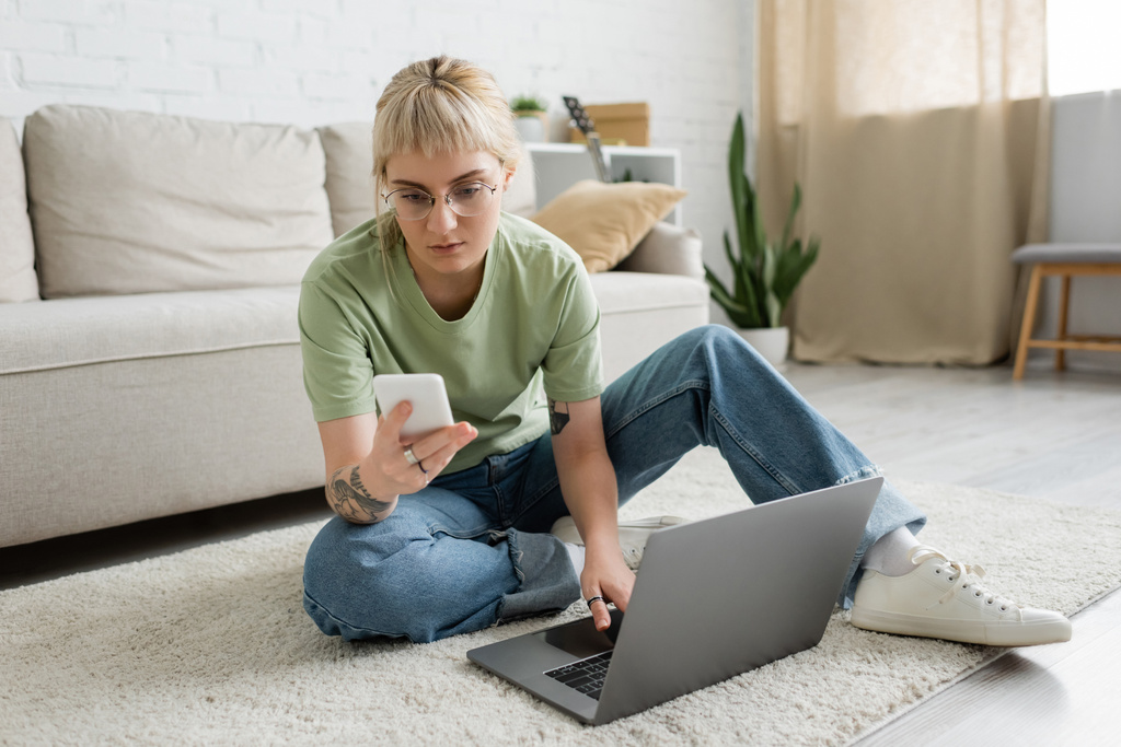 mulher tatuada com franja e óculos usando laptop enquanto sentado no tapete e segurando smartphone perto de sofá confortável e rack com plantas na sala de estar moderna  - Foto, Imagem