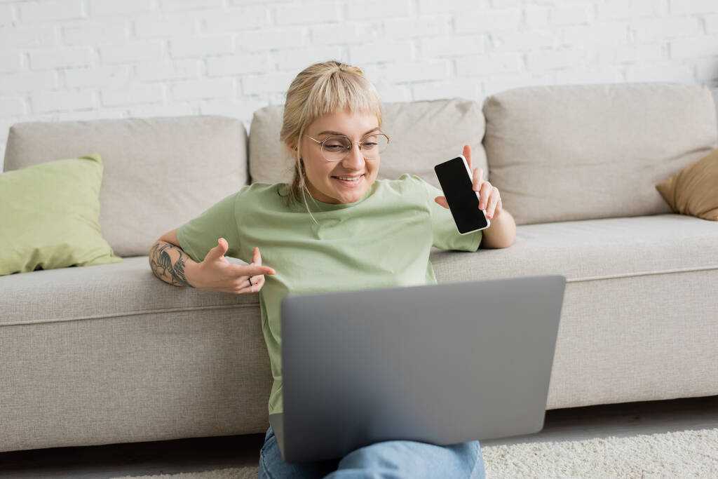 χαρούμενη και με τατουάζ γυναίκα με αφέλειες και γυαλιά που χρησιμοποιεί φορητό υπολογιστή ενώ κάθεται στο χαλί και κρατώντας smartphone με λευκή οθόνη κοντά σε άνετο καναπέ στο σύγχρονο σαλόνι  - Φωτογραφία, εικόνα
