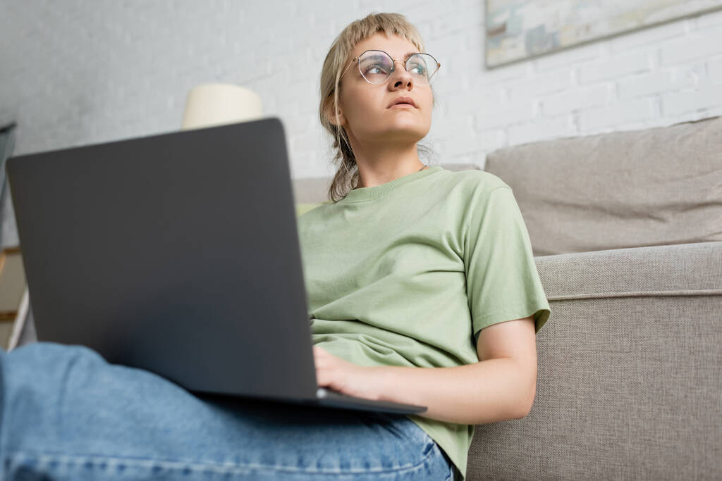 vue à angle bas de femme coûteuse avec cheveux blonds et courts, frange et lunettes à l'aide d'un ordinateur portable tout en étant assis près du canapé confortable dans le salon moderne avec paiting sur le mur  - Photo, image