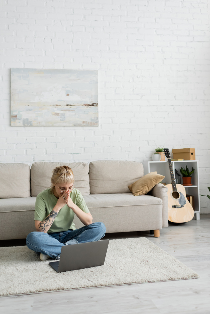татуйована жінка з чубчиками та окулярами, що закривають рот, використовуючи ноутбук, сидячи на килимі біля зручного дивана біля гітари та стійки з рослинами у сучасній вітальні з паузою на стіні
  - Фото, зображення