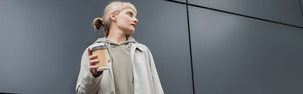 modna młoda kobieta z blond włosami z grzywką stojąca w płaszczu i kapturze trzymająca papierowy kubek z kawą na wynos w pobliżu szarego nowoczesnego budynku na ulicy, na zewnątrz, miejskiego życia, baner  - Zdjęcie, obraz