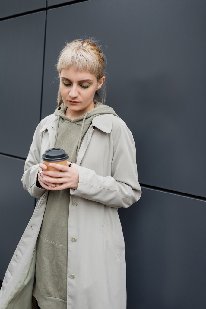 mladá žena s blond vlasy s ofinou stojící v kabátě a kapucí při držení papírového kelímku s odnášející kávou v blízkosti šedé moderní budovy na ulici, venku, městský život, street fashion  - Fotografie, Obrázek