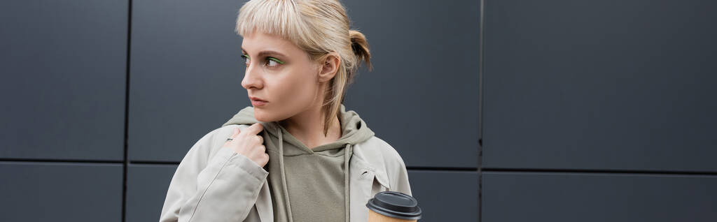 stylish young woman with blonde hair with bangs standing in coat and hoodie while holding paper cup with takeaway coffee near grey modern building on street, outside, urban living, banner  - Photo, Image