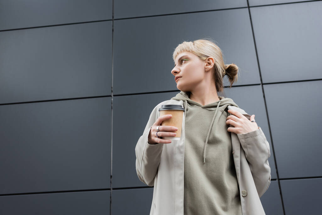 stylish young woman with blonde hair with bangs looking away and standing in coat and hoodie while holding paper cup with takeaway coffee near grey modern building on street, outside, urban living  - Photo, Image