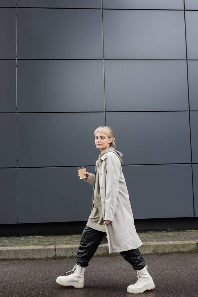 Sarı saçlı, kâküllü, ceketli, siyah deri pantolonlu, kapüşonlu ve botlu, elinde kahve bardağıyla caddedeki gri modern binanın yanında gezen modaya uygun bir kadın.  - Fotoğraf, Görsel