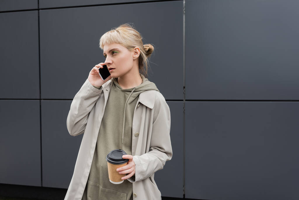 стильна молода жінка з чубчиком і світлим волоссям тримає паперову чашку з кавою, щоб піти під час розмови на смартфоні і стоячи в капюшоні і пальто біля сірої сучасної будівлі на міській вулиці
  - Фото, зображення