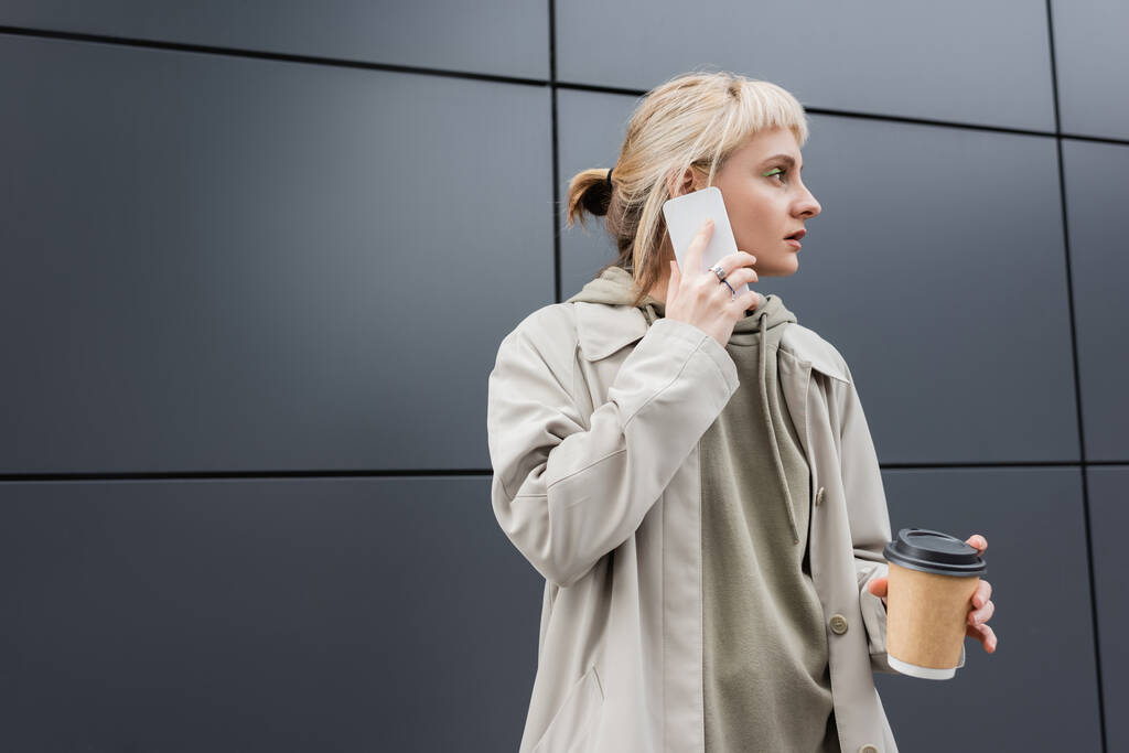 модная молодая женщина с челкой и светлые волосы держа бумажный стаканчик с кофе, чтобы пойти во время разговора на смартфоне и стоя в толстовке и пальто возле серого современного здания на городской улице, отвернуться  - Фото, изображение