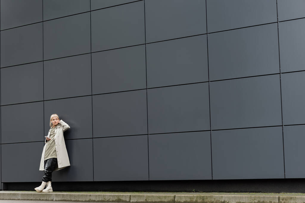 jonge vrouw met pony staan in stijlvolle kleding en draadloze oortelefoons tijdens het gebruik van smartphone in de buurt van grijs modern gebouw op stedelijke straat, jas met hoodie, zwart lederen broek en laarzen, volledige lengte - Foto, afbeelding
