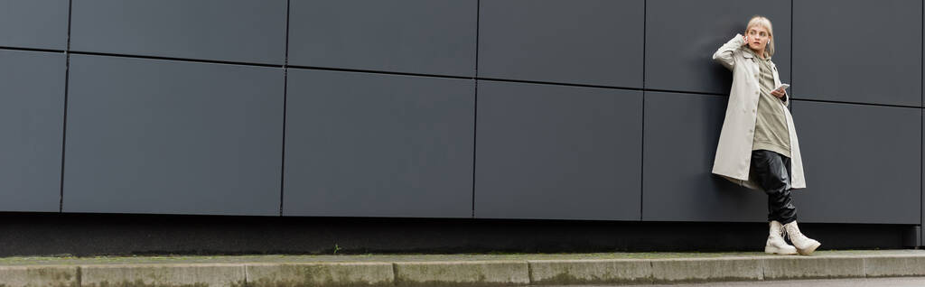 νεαρή γυναίκα με αφέλειες στέκεται σε μοντέρνα ρούχα και ασύρματα ακουστικά, ενώ χρησιμοποιώντας smartphone κοντά γκρι σύγχρονο κτίριο σε αστικό δρόμο, παλτό με κουκούλα, μαύρο δερμάτινο παντελόνι και μπότες, πανό - Φωτογραφία, εικόνα